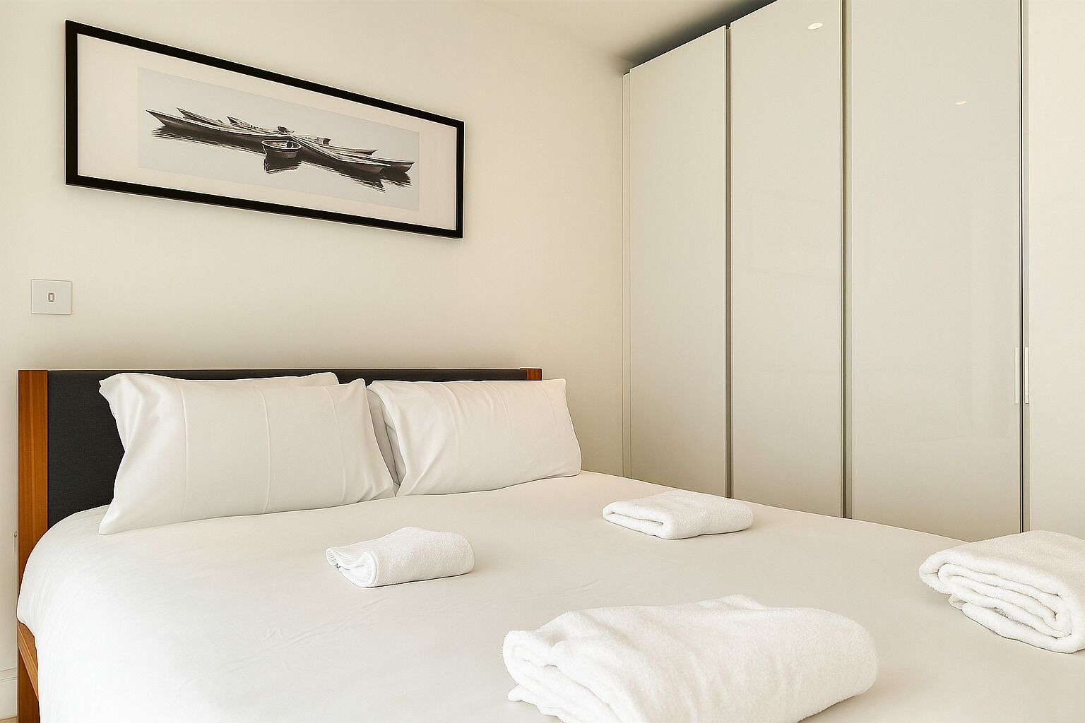 A hotel room with a bed, white pillows, a framed picture on the wall, and a wardrobe.
