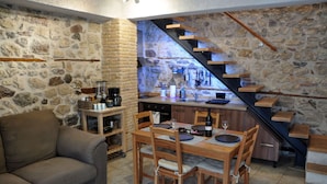 A cozy living area with a stone wall, a sofa, a dining table with chairs, and a kitchen area with a wooden staircase. - Limni Stone Apartment (Mantoudi-Limni-Agia Anna)