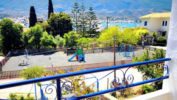 A balcony with a view of a playground, trees, and distant mountains. - Valente Perlia Rooms (Poros)