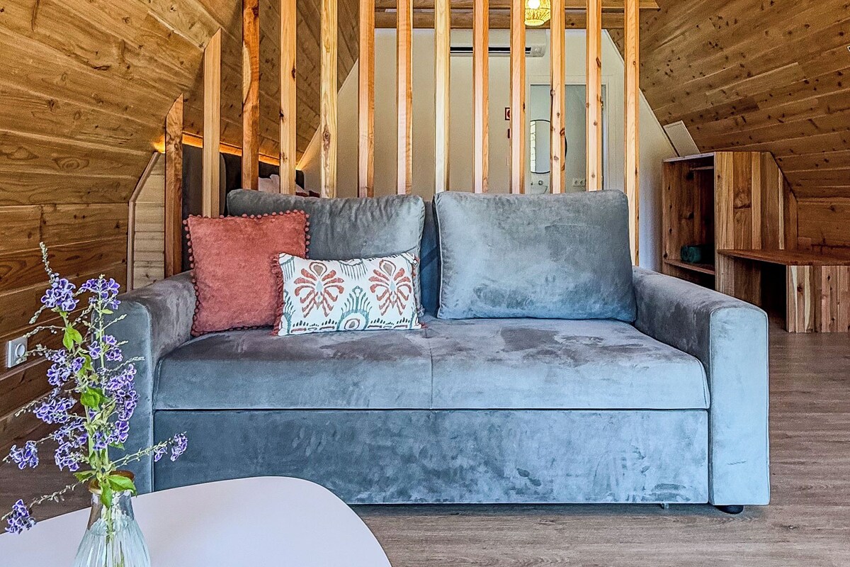 An attic room with a wooden ceiling, a grey sofa with cushions, a white table, and a vase with flowers.