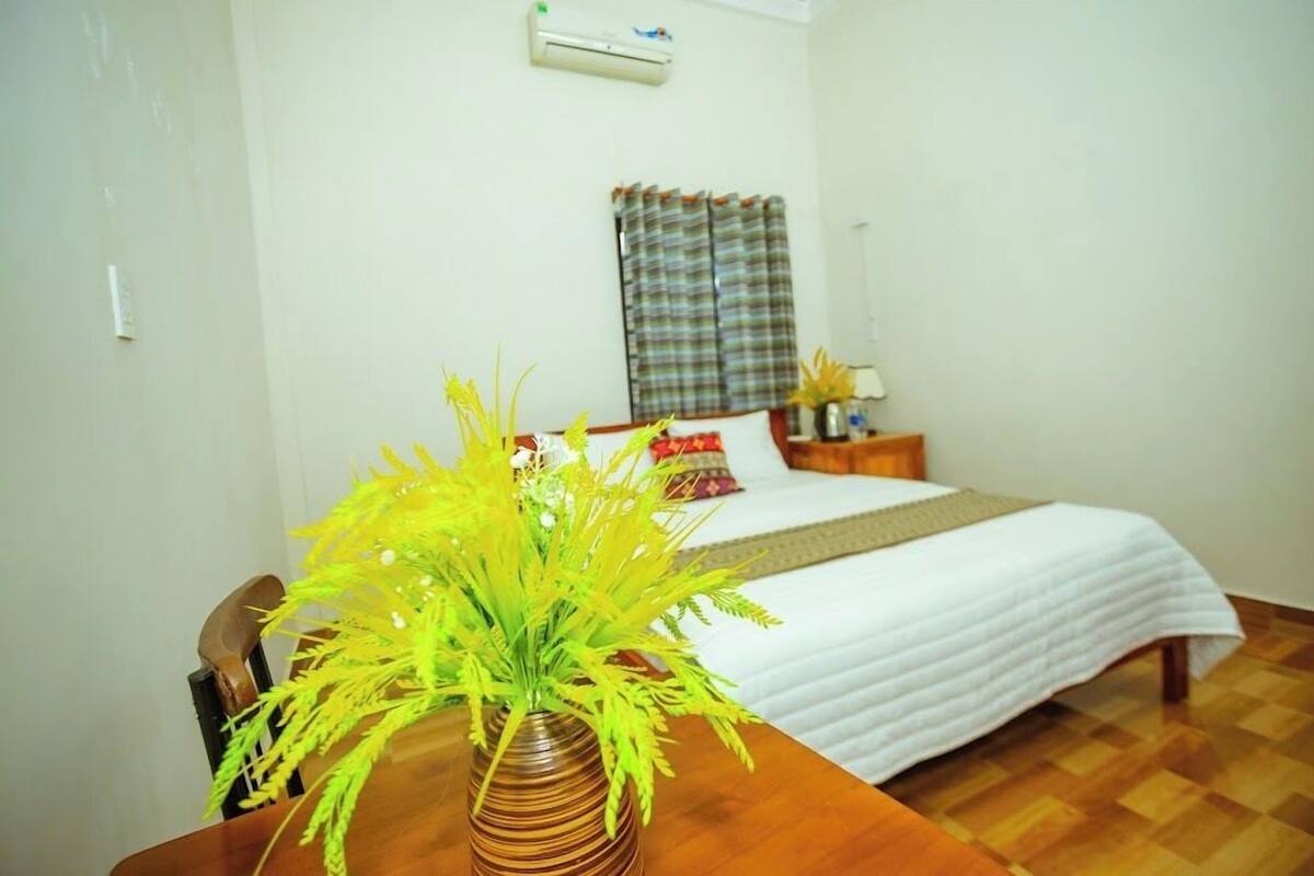 A hotel room with a bed, a bedside table with a vase of flowers, a window with curtains, and an air conditioning unit on the wall.