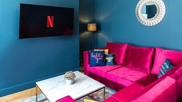 A room with a blue wall, a red sofa with cushions, a white coffee table, a TV mounted on the wall, and a floor lamp. - The Rockfield By Serene Stays (Anfield)