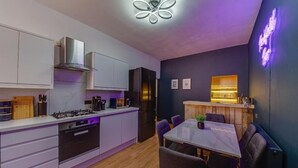 A modern kitchen with white cabinets, a gas stove, and a built-in oven. There is a dining area with a table and chairs, and a neon sign on the wall. - The Rockfield By Serene Stays (Anfield)