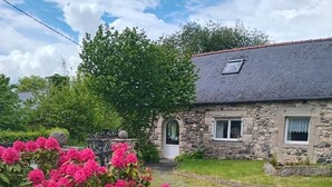 Une maison en pierre avec une allée gravillonnée, un jardin parsemé de fleurs roses et un ciel dégagé.