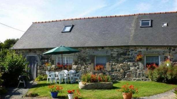 Un bâtiment en pierre avec un toit en pente, un petit porche avec des chaises blanches et des plantes en pot.