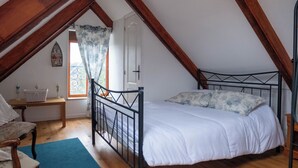 Une chambre avec un plafond en pente, un lit avec une literie blanche, une chaise en bois, une petite table et une fenêtre avec un rideau.