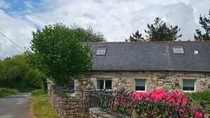 Une maison en pierre avec un toit en pente, une petite fenêtre et une clôture en métal ornée de fleurs roses.