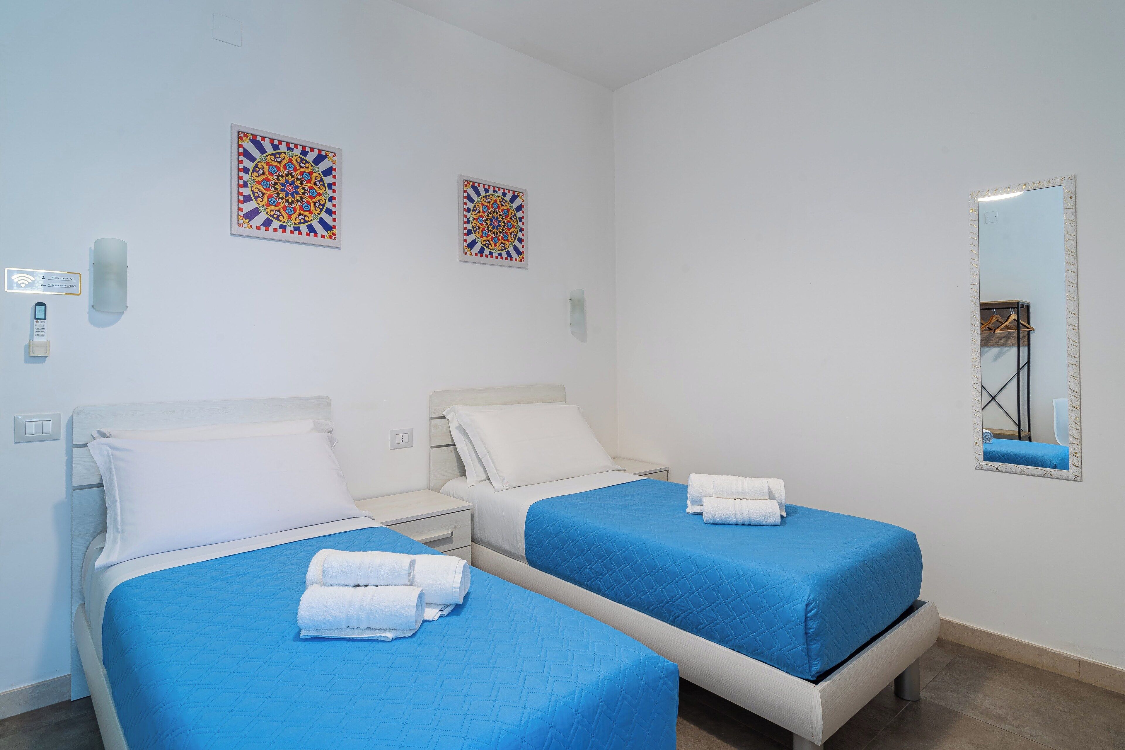 Habitació d'hotel amb dos llits, cadascun amb un llençol blau i coixins blancs, un mirall gran i dues obres d'art decoratives a la paret.
