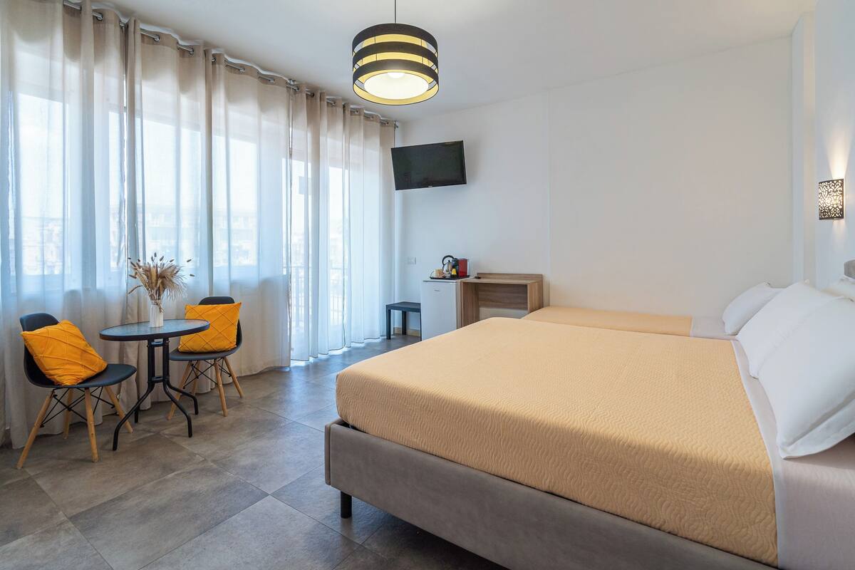 A hotel room with a large bed, a small table with two chairs, a TV mounted on the wall, and a window with curtains.