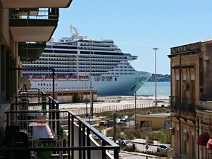 A large cruise ship docked at a port with a view of the sea and a building with balconies. - Agorà (Syracuse)