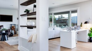 Un soggiorno moderno con un divano bianco, un tavolino da caffè e una scrivania con un laptop. Sono presenti scaffali con oggetti decorativi e un'ampia finestra che offre vista sull'esterno.