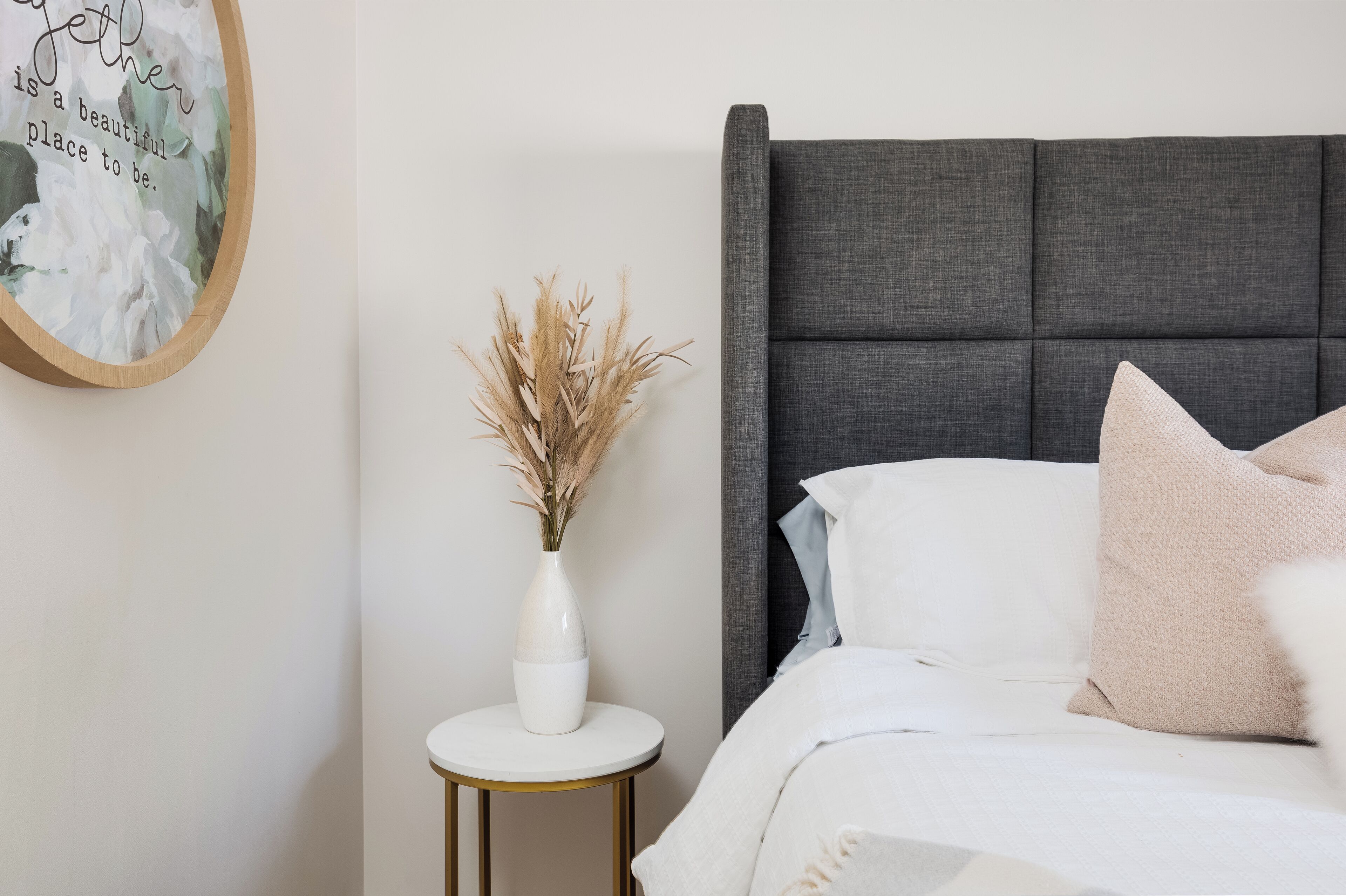 A neatly made bed with a grey upholstered headboard, a white vase with dried pampas grass on a small round table, and a framed quote on the wall.
