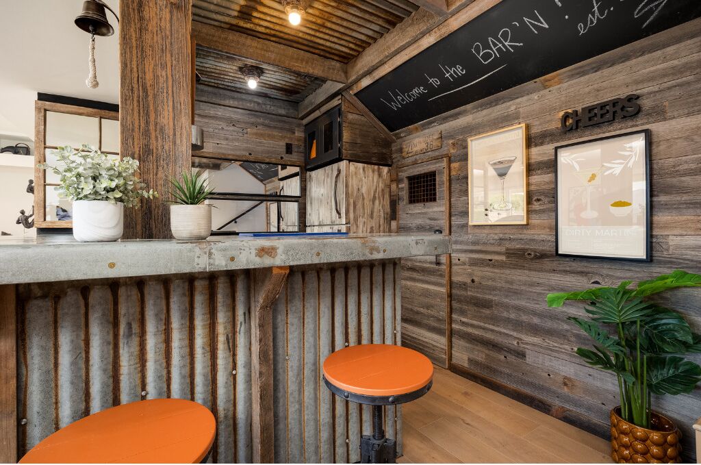 A rustic bar area with wooden walls, a chalkboard sign, framed pictures, and orange stools.