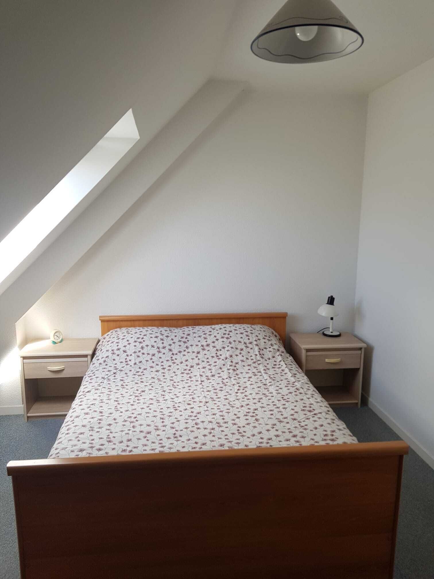 Ein Schlafzimmer mit schrägem Dach, einem Einzelbett mit floralem Überwurf, einem Nachttisch mit Lampe sowie einem kleinen Beistelltisch mit einer Uhr.