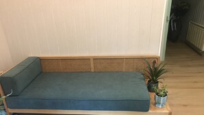 Un banc en bois avec un coussin vert, deux plantes en pot et une tête de lit en osier.