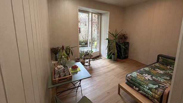 Une petite pièce comprenant un lit, une petite table avec un panier, et une fenêtre ornée de plantes.