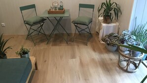 Une petite pièce avec un plancher en bois, deux chaises vertes, une petite table avec un plateau et plusieurs plantes en pot.