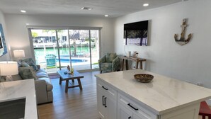 A modern kitchen with a large island, a dining area with a table and chairs, and a view of a pool and outdoor seating area through large sliding doors. - H's Hideaway - Key Colony Beach (Marathon)