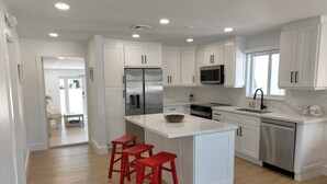 A modern kitchen with white cabinets, stainless steel appliances, a central island with stools, and a view to the adjacent living area. - H's Hideaway - Key Colony Beach (Marathon)