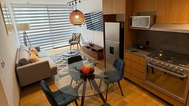 A modern kitchen with a built-in microwave, stove, and refrigerator. There is a dining area with a glass table and chairs, and a sofa in the background.