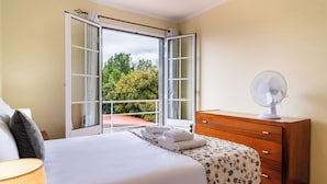 Een slaapkamer met een bed, een houten dressoir, een ventilator en uitzicht op groen via openslaande Franse deuren.