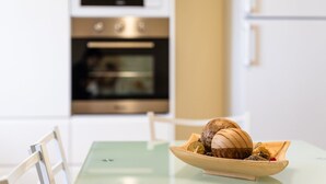 Een moderne keuken met witte kastjes, een roestvrijstalen oven en een eettafel met een glazen blad. Op tafel staat een houten schaal met een schelp en andere voorwerpen.