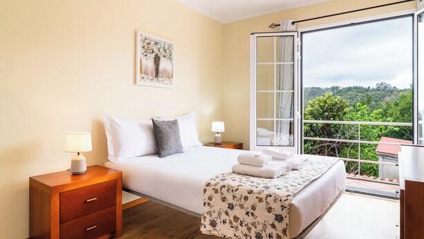 Een slaapkamer met een bed, nachtkastjes, een houten dressoir, een balkon met uitzicht op groen en een schilderij aan de muur.
