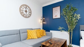 A modern living room with a grey sofa, yellow cushions, a wooden coffee table, a wall clock, a framed picture, and a potted plant. - Open Mind Property - 1 Bed Flat with 3 Double Beds (Portsmouth)