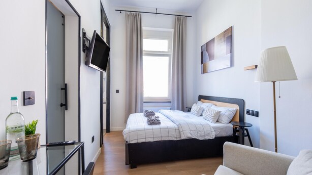 A modern hotel room with a bed, a TV mounted on the wall, a desk with a lamp, and a view of the outdoors. - Apartment in Koblenz Near Deutsches Eck (Koblenz)