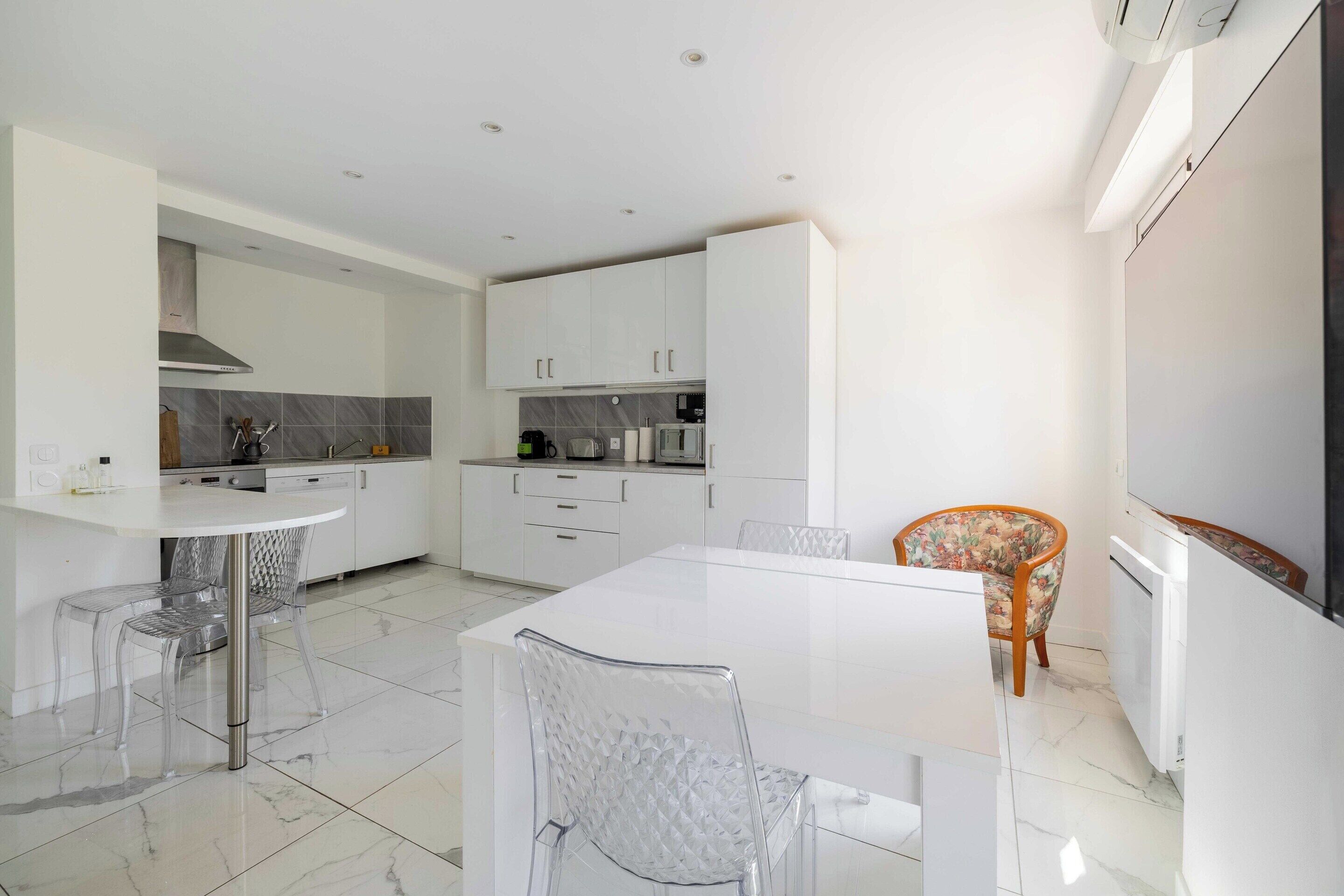 A modern kitchen with white cabinets, a white dining table, and clear chairs.