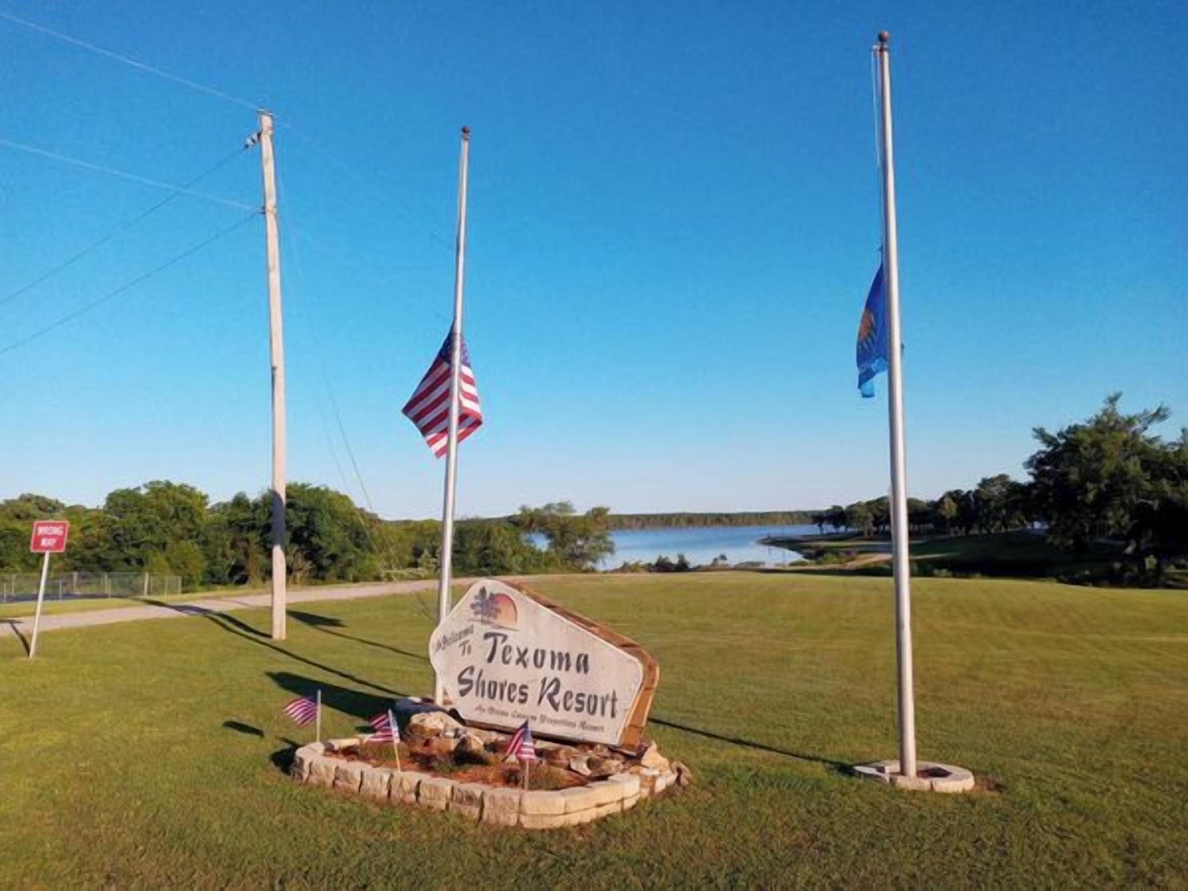 Señal de Texoma Shores Resort con banderas estadounidenses y una vista despejada de un lago al fondo.