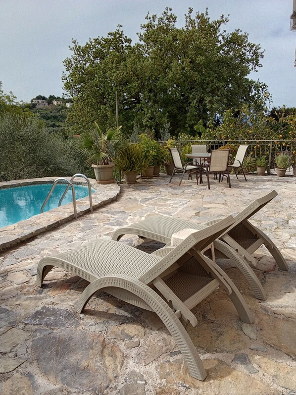 A poolside area with lounge chairs and a table, surrounded by potted plants and trees.