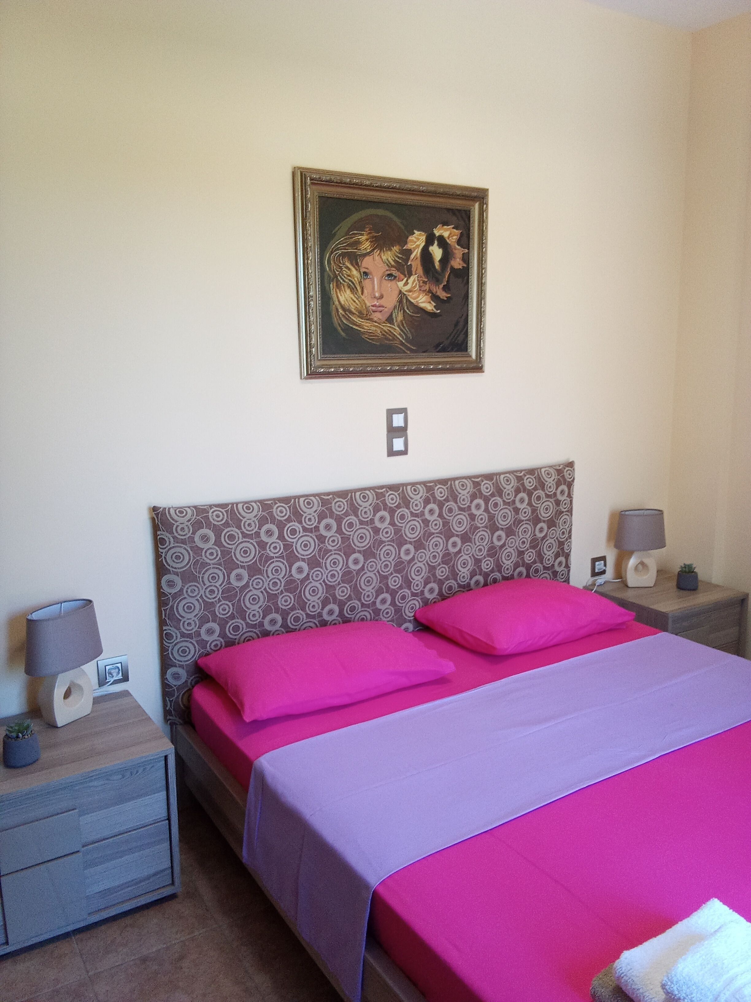 Ein Schlafzimmer mit einem gemusterten Kopfteil, einem Bett mit pinkfarbener und violetter Bettwäsche, einem Nachttisch mit Lampe und einem gerahmten Bild an der Wand.