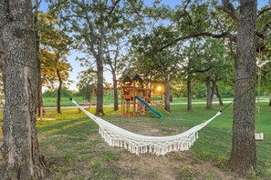 Un hamac tendu entre deux arbres dans un parc, avec une structure de jeux en bois en arrière-plan.