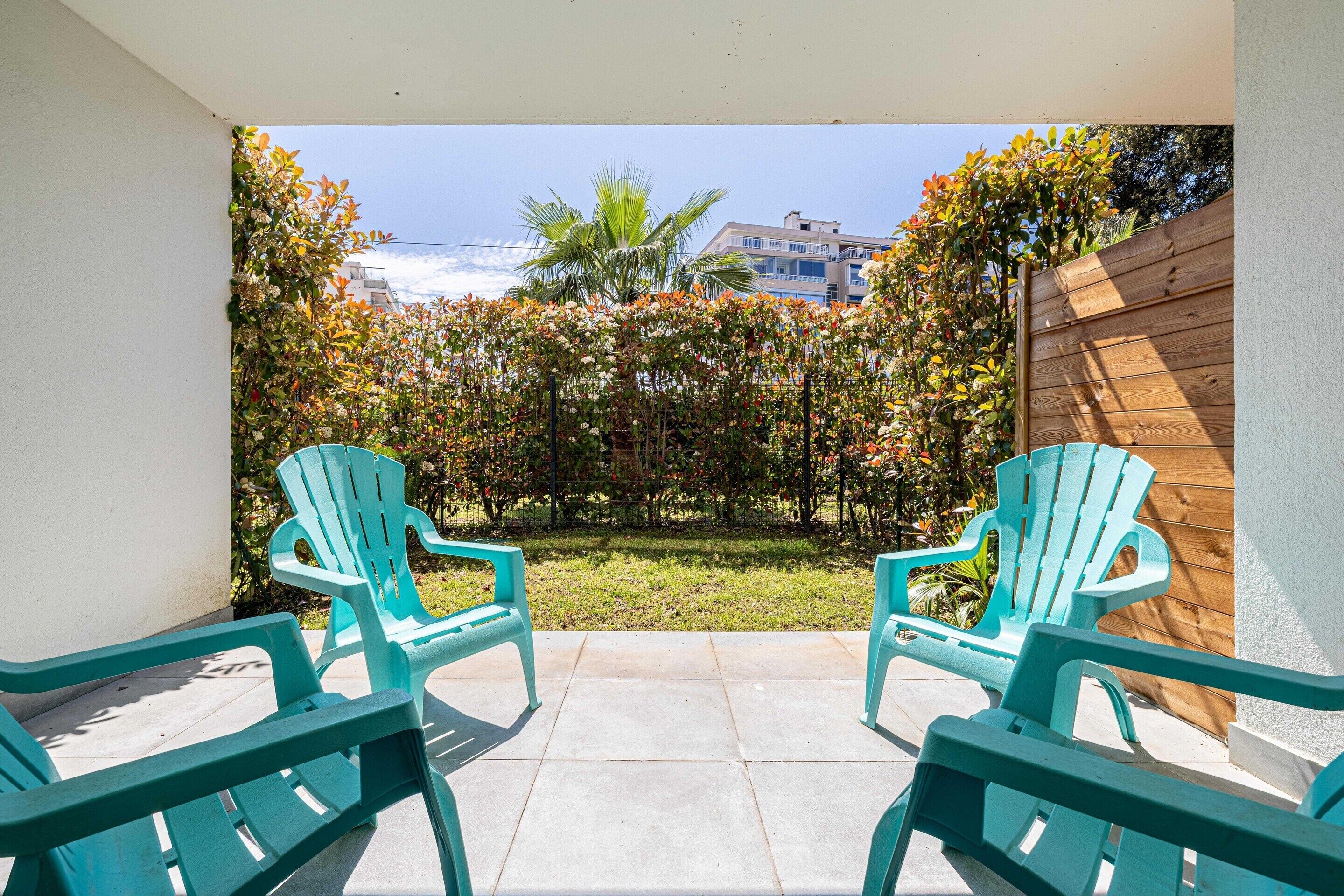 A patio with two turquoise chairs, a wooden fence, and a view of a garden with various plants and a building in the background.