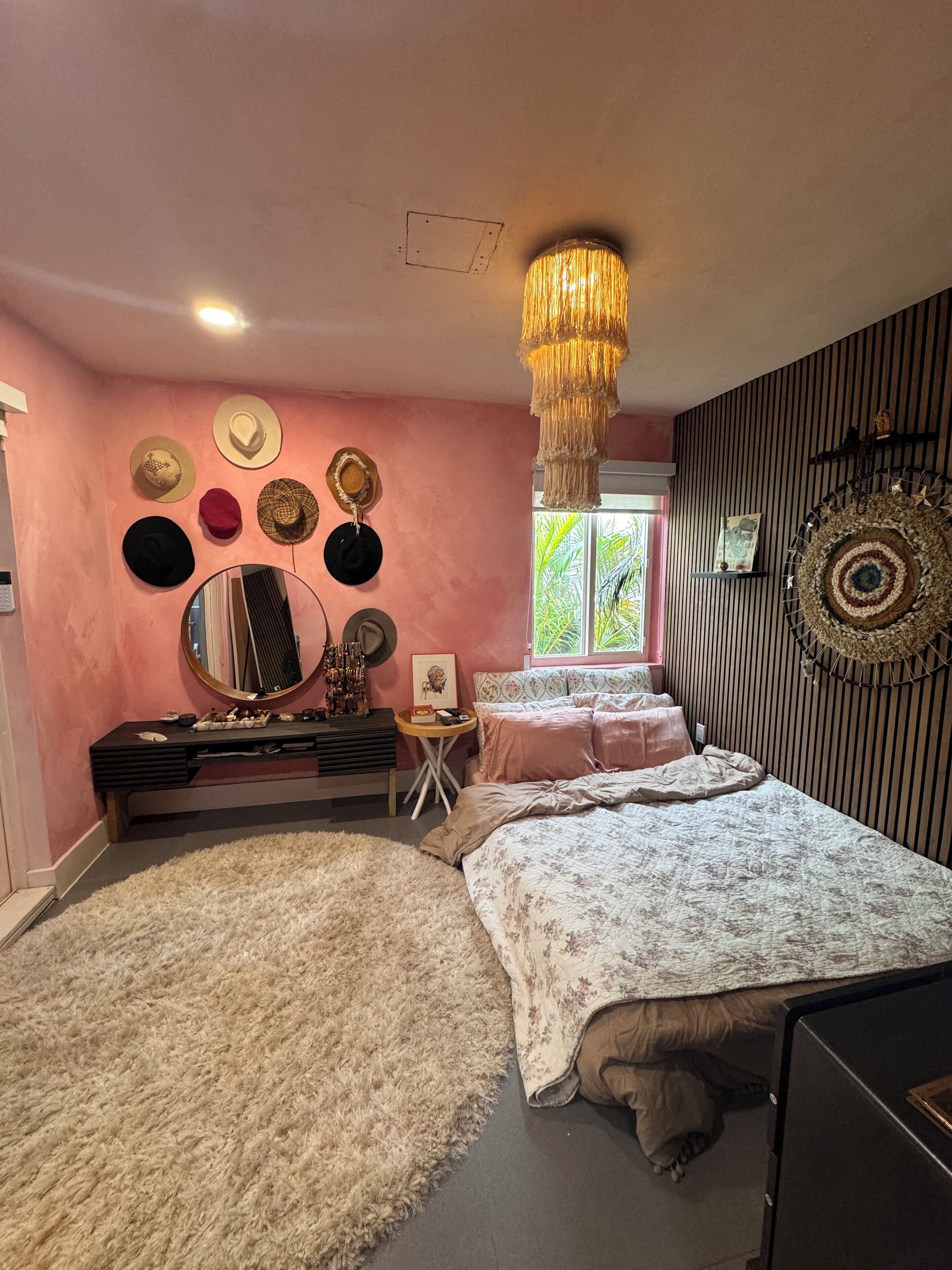 A bedroom with a bed, a vanity with a mirror, a dresser, and a wall decorated with hats and a circular mirror.