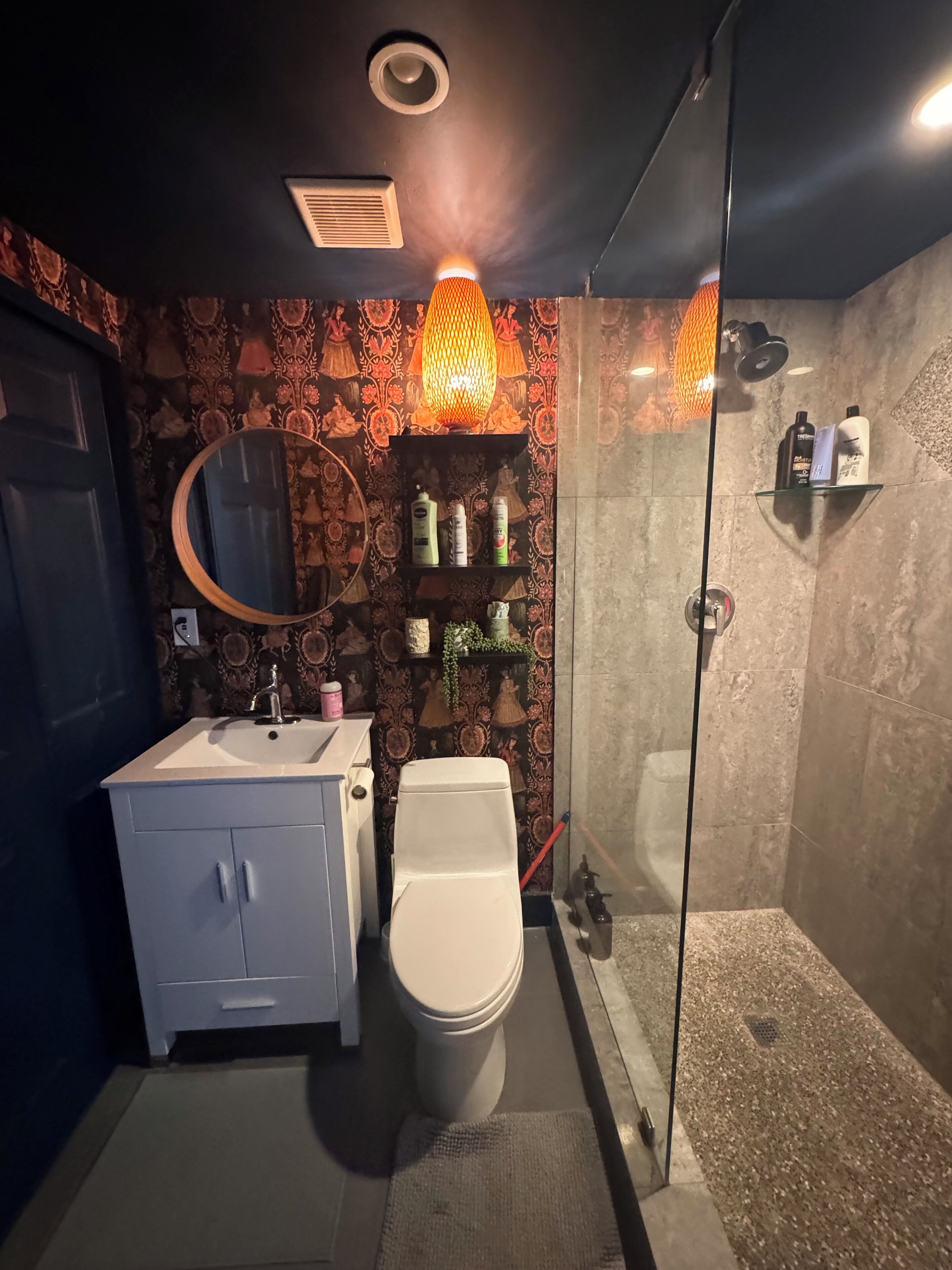 A bathroom with a toilet, sink, and shower. There is a mirror above the sink and a shelf with toiletries.