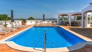 A rectangular pool with a metal safety rail, surrounded by white lounge chairs and umbrellas, with a view of distant mountains.