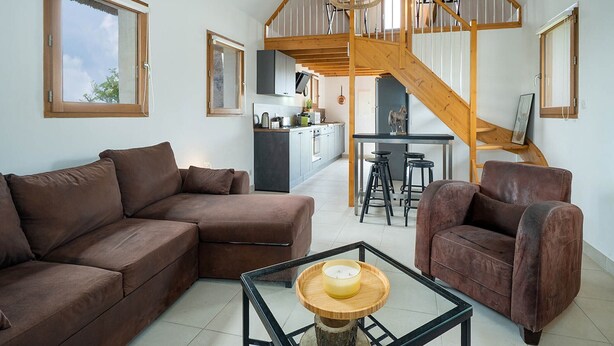 Un vaste espace de vie comprenant un canapé marron, un fauteuil marron et une table basse en verre sur laquelle est posée une bougie. Un escalier en bois mène à une cuisine équipée de tabourets de bar.