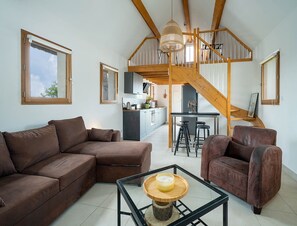 A spacious living area with a brown sofa, a brown armchair, and a glass coffee table with a candle. A wooden staircase leads to a kitchen area with bar stools.