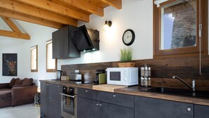 Une cuisine moderne avec des meubles de couleur sombre, un four encastré, un four à micro-ondes et un évier. Le plafond est en bois et une fenêtre offre une vue sur un mur en pierre.