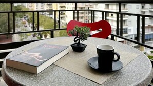 Mesa redonda con un libro, una taza negra y una pequeña planta en maceta en un balcón con una silla roja, con vistas a un paisaje urbano.