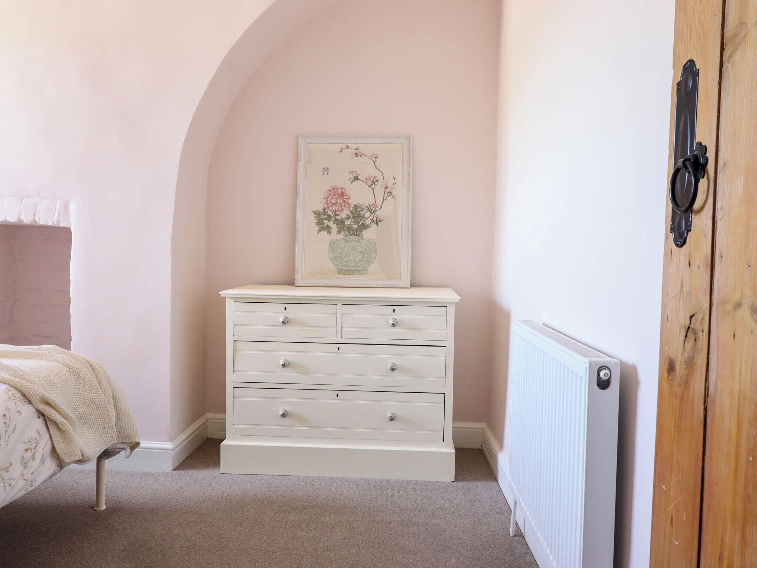 Un dormitorio con un aparador blanco, una obra de arte floral enmarco y un radiador, además de una puerta de madera.