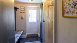 Un couloir avec une porte blanche, un banc, un porte-sac fixé au mur, une œuvre d’art florale encadrée et un luminaire au plafond.