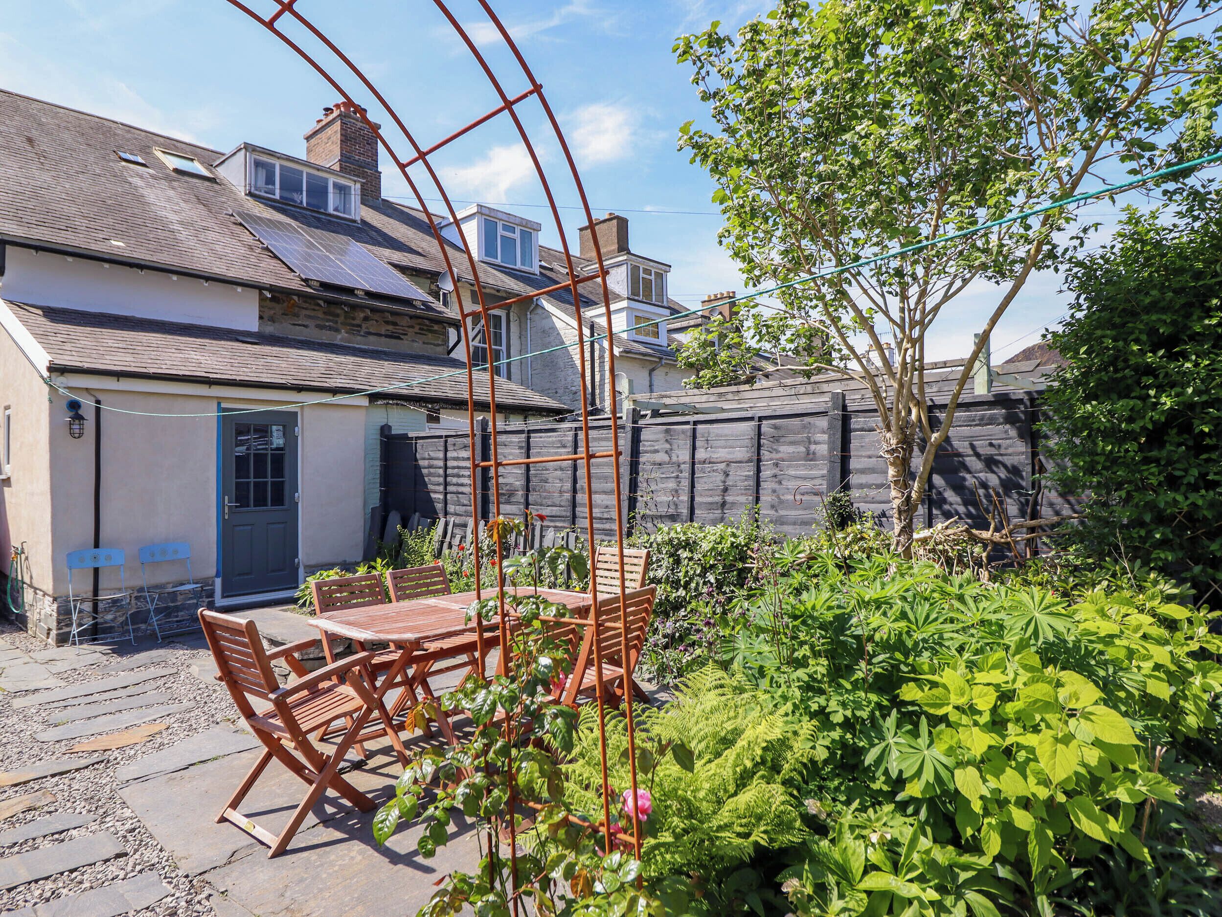 A patio with a wooden table and chairs, surrounded by lush greenery and a wooden fence.