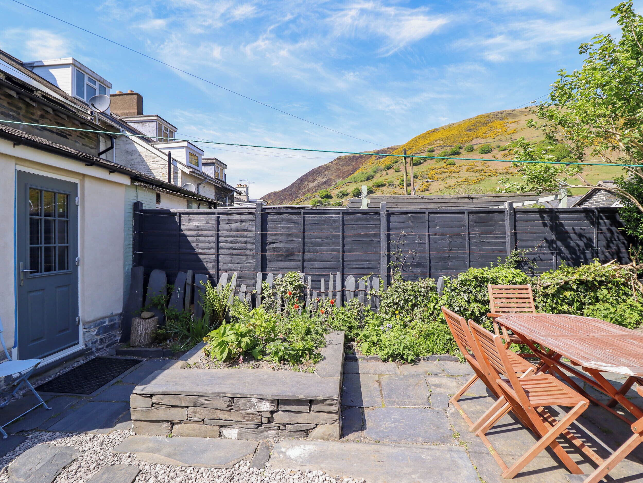 Um espaço de pátio com mesa de pedra e cadeiras de madeira, cerca preta e porta para uma casa.