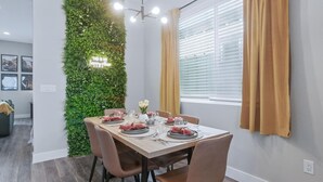 Ein Essbereich mit einem Tisch für vier Personen, umgeben von Stühlen, einer grünen vertikalen Wandgardine und einem Fenster mit Vorhängen.