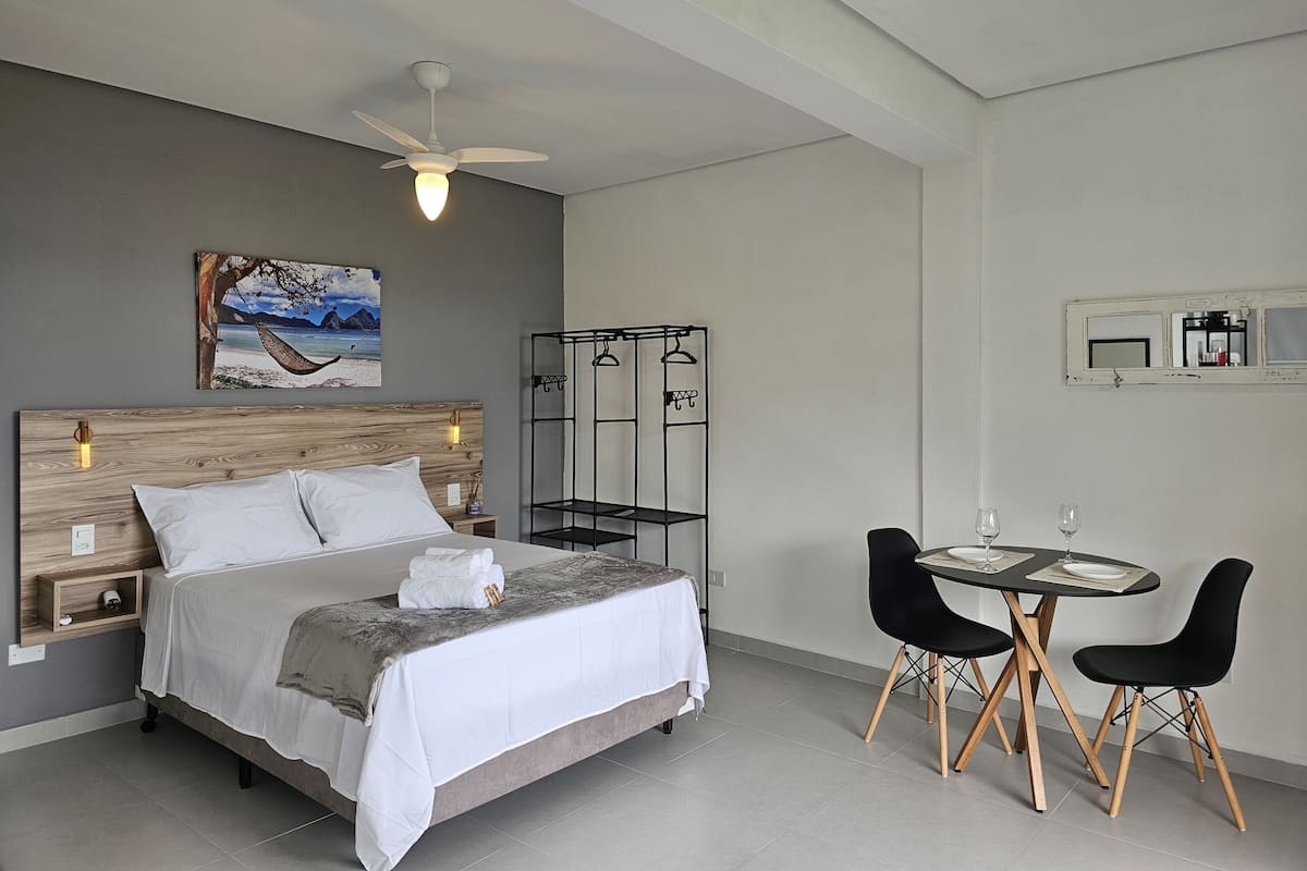 A bedroom with a bed, bedside table, nightstand, wall-mounted shelf, and a painting of a beach scene.