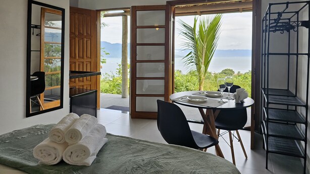 A room with a bed, a table with chairs, a view of the sea, and a wardrobe. - Suítes Yvi Porã (Ilhabela)