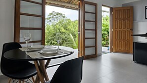 A dining area with a table set for two, a black chair, and a wooden door leading outside. - Suítes Yvi Porã (Ilhabela)